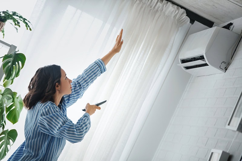 Why Is My Air Conditioner Blowing Hot Air? Woman is checking to see if the air conditioner is cooling. She is holding the remote to the air conditioner and raised her hand to check temperature.