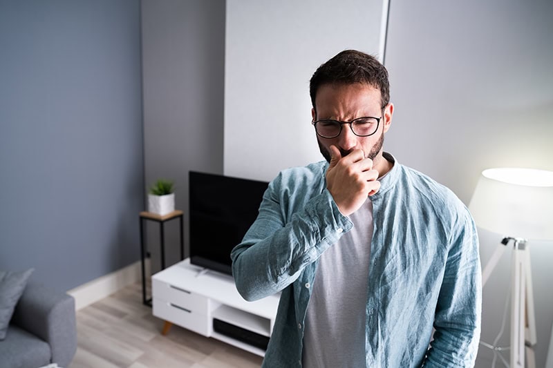 A man wearing glasses and a light blue shirt stands in a modern living room, holding a hand to his mouth as he coughs.
