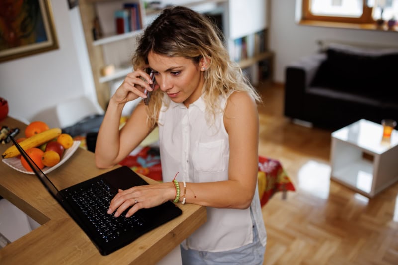 Young woman standing at kitchen counter and talking on mobile phone while working remotely on laptop from home.