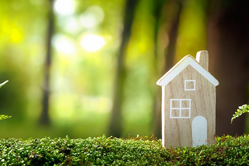 Top 5 Benefits of Installing a Geothermal System. Photo of a wooden toy house sitting on artificial grass with a blurry background of the woods.